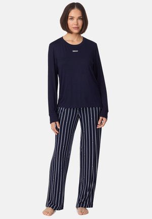 Vrouw draagt een navy blauw DKNY top met lange mouwen en marineblauwe gestreepte pyjamabroek, staat op blote voeten tegen een effen witte achtergrond.
