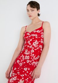 Rød blomstret kjole med hvite blomstermønstre, tettsittende design, tynne stropper og en thigh-high splitt på den ene siden, laget av lett stoff.