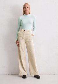 Hellgrünes Langarmoberteil kombiniert mit beigen, weiten Cordhosen. Bemerkenswerte Details sind die vorderen Taschen und ein einzelner Knopfverschluss.