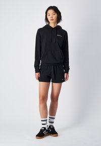 Czarna bluza z kapturem i kieszenią z przodu, zestawiona z czarnymi szortami sportowymi. Posiada haftowane logo zarówno na bluzie, jak i szortach. Czarne sneakersy i białe skarpetki w paski.