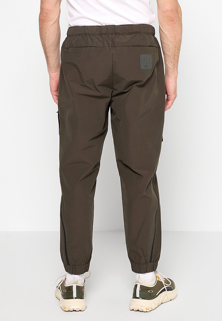 Uomo che indossa pantaloni jogger elastici verde oliva con caviglie a costura e due tasche posteriori, abbinati a calzini bianchi e scarpe da ginnastica verdi.