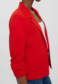 Punainen bleiseri, jossa on ryhdikäs kaulus ja kolme neljänneksen mittaiset hihat. Siinä on sileä pinta ja mittatilaustyönä tehty istuvuus. Ei näkyviä koristeita.