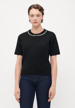Young woman with short hair wearing a black short-sleeve sweater decorated with a circle of white beads around the neckline and dark jeans.