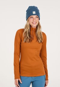 Langsleeve, hoge hals top in warme oranje stof, gecombineerd met een blauwe geribde beanie met een logo patch. Soepele textuur, figure-hugging ontwerp.