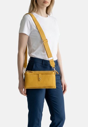 Donna che indossa una maglietta bianca e pantaloni blu navy con una borsa a tracolla in pelle giallo senape con tracolla larga regolabile.