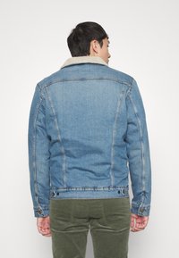 Denims jaka ar ādas apdari, gaiši zila krāsa, klasiskā dizainā un redzamām šuvēm. Aizmugures skats rāda strukturētu piegriezumu.