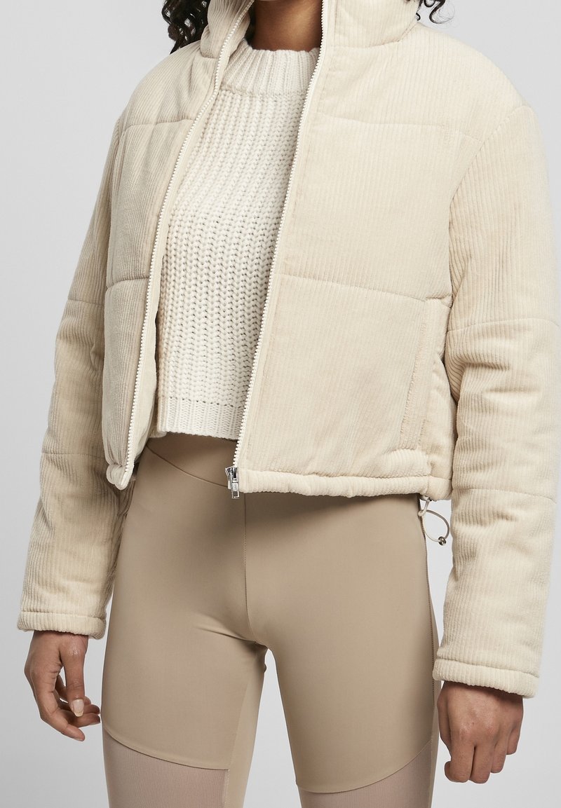 Personne portant une doudoune en velours côtelé beige par-dessus un pull en maille blanc et un pantalon beige ajusté, debout devant un fond uni.