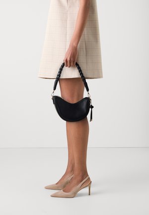 Borsa a tracolla in pelle nera con forma curva, manico intrecciato e chiusura con zip, tenuta in mano vicino a un abito a quadri fino al ginocchio e tacchi nudi.