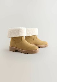 Ljusbruna mocka ankelskor med en vit faux fur-mudd, gummiyttersula och sömnad längs sulan. Rund tådesign.