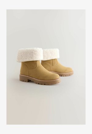 Tan suède enkellaarzen met een witte nepbont manchet, rubberen buitenzool en stikselaccenten langs de zool. Ronde neusdesign.