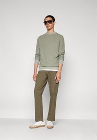 Hellgrüner Pullover mit gerippten Bündchen, über einem weißen Hemd getragen; kombiniert mit olivgrünen Hosen und off-white Sneakers mit beigen Akzenten.