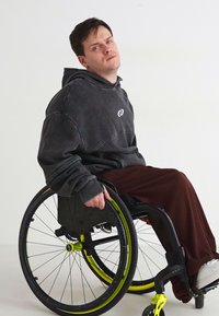 Mustaga kapuutsiga sviiter, millel on rindkeres logo, koos laiemate pruunide pükstega. Isik istub musta ja kollase disainiga sujuva ratastooliga.