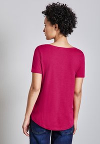 T-shirt a maniche corte e collo rotondo in tessuto rosa brillante, contaglio dritto e orlo leggermente curvo, abbinato a jeans blu scuro.
