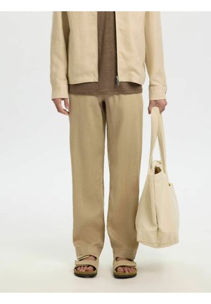 Person trägt beige Hose, beige Jacke, braunes Hemd, beige Sandalen und hält eine beige Tragetasche an den Griffen an der Seite.