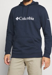 Man i marinblå Columbia-tröja med framficka och beige byxor, som står mot en enfärgad bakgrund.