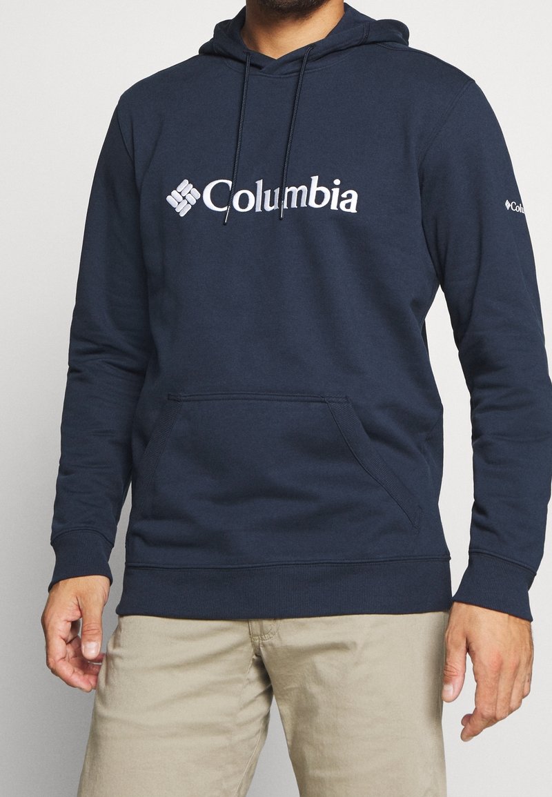 Man i marinblå Columbia-tröja med framficka och beige byxor, som står mot en enfärgad bakgrund.