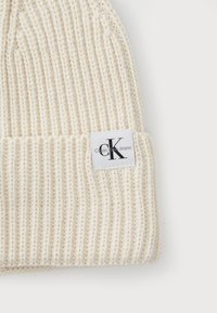 Krémová pletená čapica s ribbovanou textúrou a preloženým golierom. Obsahuje bielu nálepku s čiernym logom "Calvin Klein Jeans".