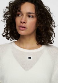 Maglione bianco in rete con design a V sul collo, dettaglio del logo quadrato nero sul petto e un tessuto testurizzato e traspirante.