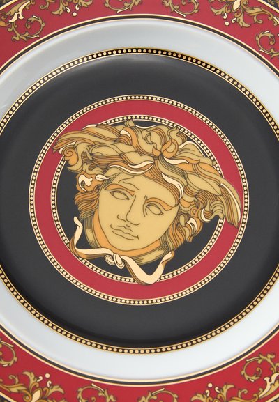 Okrągły talerz z centralnym złoto-czarnym medalionem przedstawiającym stylizowaną twarz, otoczonym czerwonym obrzeżem z misternymi złotymi zdobieniami.