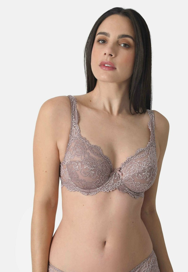 Woman wearing mauve lace bra with floral patterns, standing against a plain light background, facing forward with neutral expression.