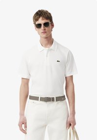 Camisa polo de algodón blanca con cuello clásico, mangas cortas y un logo bordado verde en el pecho. Combinada con pantalones blancos y un cinturón gris.