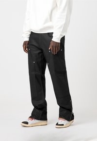 Pantalon cargo noir avec multiples poches, fermetures éclair argentées aux chevilles, porté avec un sweat à capuche blanc et des baskets multicolores.