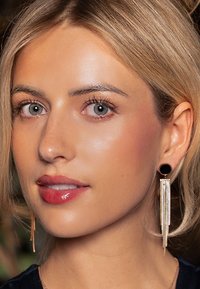 Des boucles d'oreilles pendantes dorées présentent un accent circulaire noir en haut et des franges métalliques en cascade. Le modèle a des cheveux blonds clairs et des lèvres brillantes.