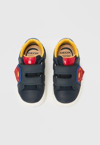 Sötétkék tornacipők sárga béléssel, gumitalppal, és két tépőzáras pánttal, melyeken oldalról piros-kék szuperhős embléma látható.