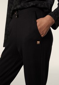 Pantaloni da jogging neri realizzati in tessuto morbido, con una vita elasticizzata, tasche laterali e un accento con logo in tono oro sull'anca sinistra.