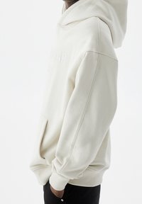 Persona che indossa un hoodie bianco sporco con testo in rilievo e mani nella tasca frontale, mostrata di lato su uno sfondo bianco semplice.