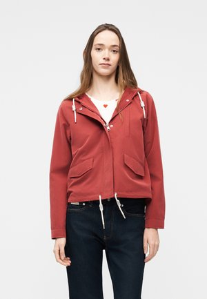 Jeune femme portant une veste à capuche rouge sur un t-shirt blanc avec un cœur rouge, associée à un jean bleu foncé, debout devant un fond uni.