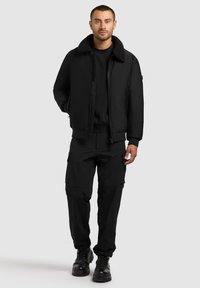 Jachetă bomber neagră cu guler din blană sintetică, purtată cu pantaloni cargo negri și bocanci negri cu șireturi. Culoare solidă, design casual, buzunare funcționale.