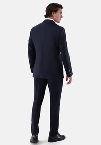 Marineblauer Anzug mit tailliertem Schnitt, einreihigem Sakko und flachen Vorderhosen, kombiniert mit schwarzen Schuhen. Glatte Stoffstruktur.