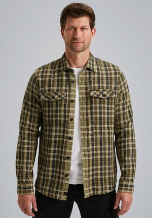 Camisa de franela a cuadros verde y negra con cierre de botones, dos bolsillos en el pecho y mangas largas. Llevada sobre una camisa blanca.
