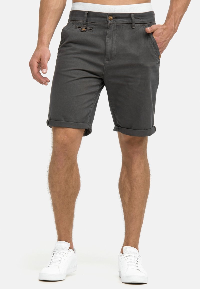 Homme portant un short gris foncé avec ourlets roulés, un sous-vêtement à taille blanche et des baskets blanches, debout devant un fond uni.