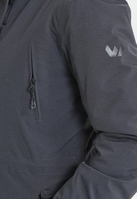 Veste gris foncé avec une poche zippée sur la manche gauche, d'une texture lisse et d'un logo discret sur l'épaule.