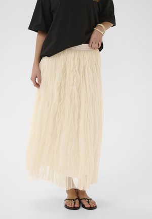 Ženska z oblečeno teksturirano kremno maksi krilo, črno preveliko majico, sandali in zapestnicami, stoji ob enostavni ozadju.