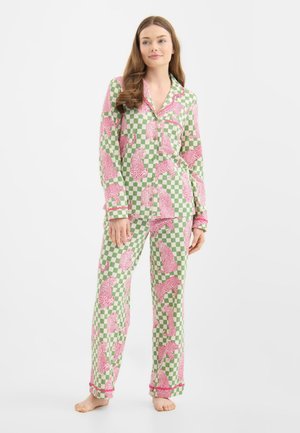 Donna che indossa un pigiama a quadri verdi e bianchi con motivi di leopardo rosa, in piedi a piedi nudi su uno sfondo piatto.