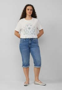T-shirt blanc avec un motif floral vert, associé à un jean bleu à revers et des ballerines argentées, se tenant devant un fond neutre.