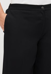 Svarte bukser laget av glatt stoff, med knappelukking, sidelommer og flat frontdesign. Enkle uten synlige mønstre.