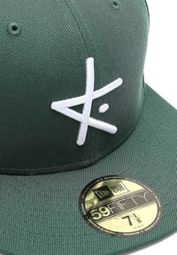 Grüne Stoffmütze mit einem weißen gestickten Logo, flachem Schirm und einem runden New Era-Aufkleber auf der Vorderseite. Sichtbare strukturierte Oberflächenbeschaffenheit.