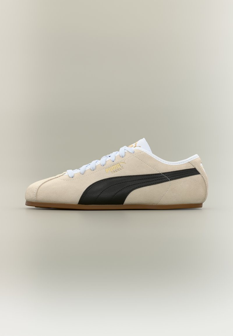 Beige Puma semsket skinnsneaker med hvite snøringer, svart sidestripe, gummisåle og gullfarget Puma-logo på nøytral bakgrunn.