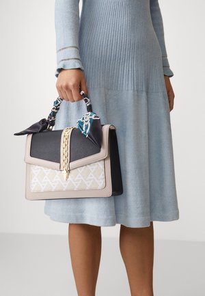 Femme en robe côtelée bleu clair tenant un sac à main beige, noir et blanc avec une chaîne dorée et un foulard à motifs enroulé autour de la poignée.