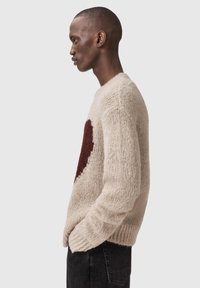 Μπεζ πλεκτό πουλόβερ με μεγάλο μοτίβο σκούρου κόκκινου καρδιού στο μπροστινό μέρος. Η υφή είναι απαλή και χοντρή, με ριμπ μανσέτες και τελείωμα.