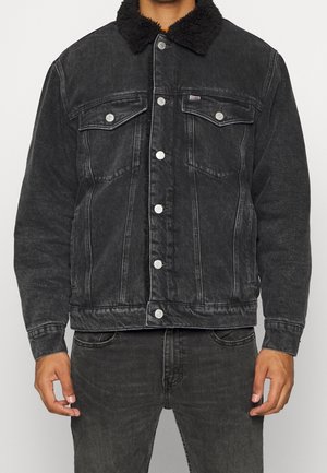 Schwarze Denimjacke mit Lammfellkragen, vorne mit Knöpfen, Klappentaschen auf der Brust, getragen von einer Person in passenden schwarzen Jeans.