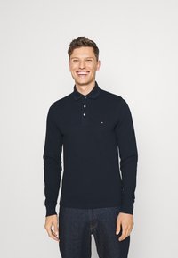 Polo à manches longues bleu marine en coton, doté d'une patte de boutonnage à deux boutons et d'un petit logo sur la poitrine. Porté avec un jean foncé.