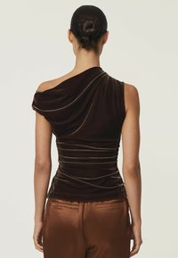 Bruine fluwelen top met één schouder, met gedrapeerd materiaal, geplooide details langs de taille en een zachte textuur, gedragen met bruine satijnen broeken.