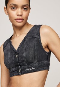 Svart denim crop top med V-hals, frontknapper og brodert "Tommy Jeans" logo på kanten. Har en tettsittende design og er ermeløs.