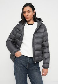 Γκρι μπουφάν puffer με κουκούλα, με ραμμένο σχέδιο, εμπρόσθιο φερμουάρ και ελαστικά μανίκια, φορεμένο πάνω από λευκό πουλόβερ και μπλε τζιν.