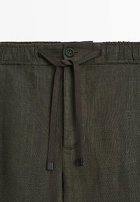 Linen blend trousers in dark green with an elastic waistband, featuring a drawstring closure and a button accent. Texture is slightly textured.